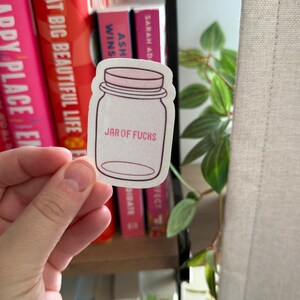 Op de afbeelding: Een sticker in de vorm van een weckpot met de woorden "JAR OF FUCKS" erop gedrukt. De sticker wordt voor een boekenplank met boeken in verschillende kleuren gehouden.