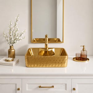 May include: A rectangular gold bathroom sink with a textured geometric honeycomb pattern on the exterior, paired with a matching gold faucet, a gold vase with white flowers, a white towel with soap, and a gold soap dispenser set.