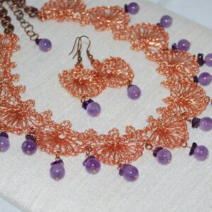 Pode incluir: Um conjunto de colar e brincos de fio de cobre com pedras de ametista roxa. O colar apresenta um design delicado e intrincado, enquanto os brincos são simples e elegantes.