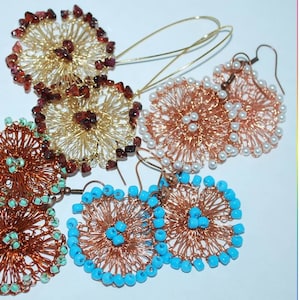 Könnte beinhalten: Eine Sammlung handgefertigter Drahtohrringe in verschiedenen Farben und Designs. Einige Ohrringe haben Golddraht mit roten Steinen, andere Kupferdraht mit türkisfarbenen Perlen und einige Kupferdraht mit weißen Perlen. Die Ohrringe sind auf weißem Hintergrund ausgestellt.