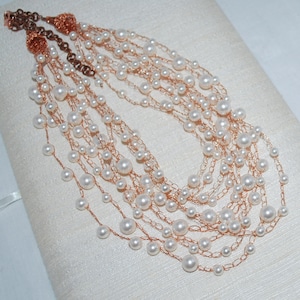 Pode incluir: Um colar de várias camadas feito de fio de cobre e pérolas falsas brancas. O colar é em camadas e apresenta uma variedade de tamanhos de pérolas.