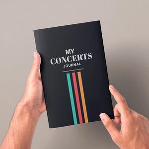 May include: A black rectangular journal with the white text MY CONCERTS JOURNAL printed on the front cover. Below the text are three vertical stripes in teal, red, and orange, extending from a horizontal line toward the bottom edge of the cover.