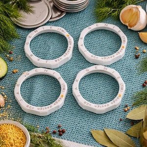 Set of four white Jar Genie magic screw rings arranged on a blue towel with canning ingredients like garlic and peppercorns.