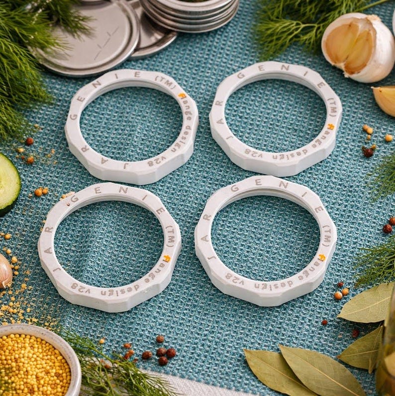 Set of four white Jar Genie magic screw rings arranged on a blue towel with canning ingredients like garlic and peppercorns.