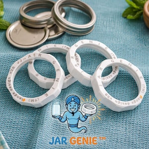 May include: White jar rings with the text "Range design v28" and "JAR GENIE" logo. Several silver metal jar lids and rings are in the background. The items are on a blue textured surface.