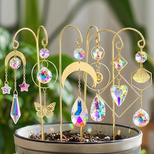 Könnte beinhalten: Ein Set aus sechs goldfarbenen Metall-Pflanzensteckern mit hängenden Ornamenten. Jeder Stecker zeigt verschiedene geometrische Formen wie Monde, Sterne, Herzen und Schmetterlinge, verziert mit facettierten, irisierenden Glaskristallen.