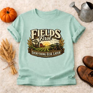 May include: A light blue t-shirt with a farm-themed graphic. The graphic includes the text "FIELDS First EVERYTHING ELSE LATER" with a tractor, barn, and wheat. A pumpkin, wheat stalks, and a watering can are also visible.