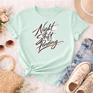 Puede incluir: Una camiseta verde claro con mangas enrolladas. La camiseta presenta el texto "Night Shift Pending" en una elegante escritura negra y dorada. La camiseta está anudada a la cintura y se combina con pantalones cortos de mezclilla y zapatillas blancas.