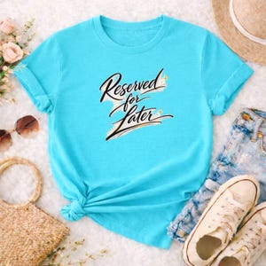 Pode incluir: Uma t-shirt azul clara com mangas enroladas e a frase "Reserved for Later" em um script elegante. A t-shirt é combinada com calções de ganga e tênis brancos, sugerindo um estilo casual e descontraído.