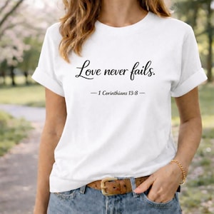 Könnte beinhalten: Weißes T-Shirt mit dem Text "Love never fails." in kursiver Schrift, gefolgt von "- 1 Korinther 13:8 -". Das Shirt hat hochgekrempelte Ärmel und wird mit Jeans und einem braunen Gürtel kombiniert.