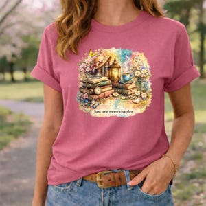 Puede incluir: Camiseta rosa con una colorida ilustración de libros, flores, una linterna y una taza de té. El texto "Just one more chapter" está debajo. La camiseta tiene mangas enrolladas.