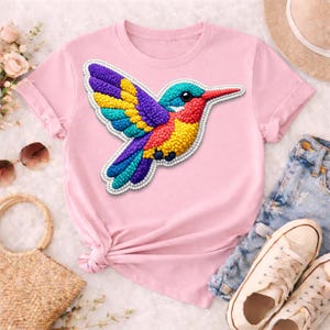 May include: A light pink t-shirt featuring a colorful hummingbird design. The hummingbird is depicted with vibrant hues of blue, purple, yellow, and red, with a detailed beaded texture. The shirt is styled with rolled-up sleeves and a knotted front.