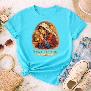 Pode incluir: Camiseta azul claro com uma ilustração religiosa de uma mulher rezando, com o texto dourado "PRAYER HEARD". A mulher usa um véu branco e uma túnica vermelha e azul. O fundo inclui velas e um brilho quente.