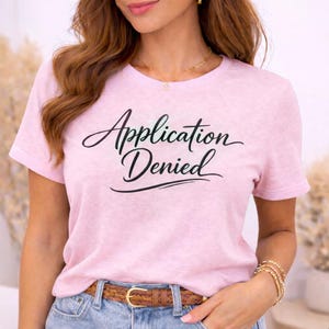 May include: A light pink t-shirt with the words "Application Denied" in black script. The shirt has short sleeves and a crew neck. The model is wearing blue jeans and a brown belt.