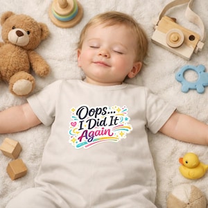 Puede incluir: Un body de bebé color crema con el texto "Oops... I Did It Again" en un diseño colorido. El bebé está acostado sobre una manta blanca, rodeado de juguetes, incluyendo un oso de peluche y una cámara de madera.