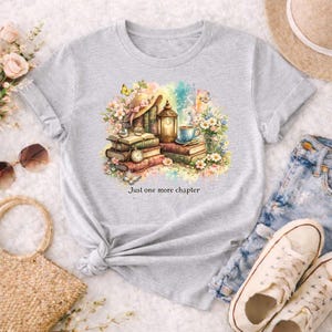 Puede incluir: Camiseta gris claro con un diseño de acuarela de libros, una taza de té, una linterna y flores. El diseño incluye el texto "Just one more chapter". La camiseta es una prenda casual para el día a día.
