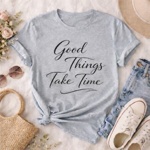 Könnte beinhalten: Hellblaues T-Shirt mit dem Text "Good Things Take Time" in schwarzer Schrift. Das Shirt hat hochgekrempelte Ärmel und einen geknoteten Bund. Accessoires sind Sonnenbrille, Strohtasche und weiße Turnschuhe.