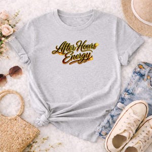 May include: A light gray t-shirt with the text "After Hours Energy" in a gold and red script. The shirt is styled with rolled-up sleeves and a knot at the waist. Accessories include sunglasses, a straw bag, a hat, and sneakers.