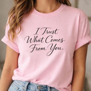 Pode incluir: T-shirt rosa claro com o texto preto "I Trust What Comes From You". As mangas estão enroladas. A t-shirt é feita de um material macio.