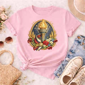 Pode incluir: Uma t-shirt rosa claro com uma ilustração detalhada de uma tocha com uma chama, rodeada por trigo, flores e uma fita em vermelho, branco e azul. O design é colocado sobre um fundo azul escuro com detalhes dourados.
