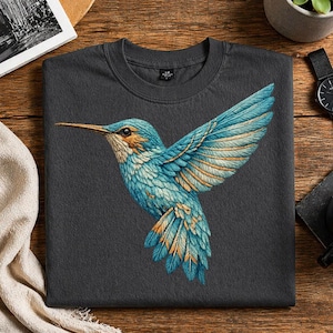 Puede incluir: Camiseta gris oscuro de cuello redondo con una ilustración detallada de un colibrí en vuelo. El ave tiene plumas azules, turquesas y doradas, con un pico largo. El diseño está centrado en la camiseta, creando un efecto visual llamativo.