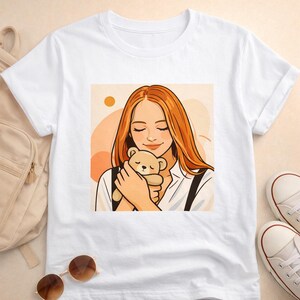 Könnte beinhalten: Weißes T-Shirt mit einer Cartoon-Illustration einer Person mit langen roten Haaren, die einen Teddybären hält. Die Illustration verwendet Orange- und Beigetöne. Das Shirt hat einen klassischen Rundhalsausschnitt und kurze Ärmel.