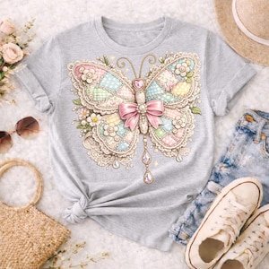 May include: A light gray t-shirt featuring a detailed butterfly design. The butterfly has patchwork wings in pastel colors, a pink bow, and dangling crystal accents. The design is embellished with floral and lace details.
