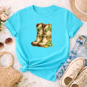 Puede incluir: Una camiseta azul claro con un diseño de botas de vaquero adornadas con tréboles, monedas de oro y hebillas en forma de corazón. Las botas son de color beige y crema, con tréboles verdes y detalles dorados. La camiseta tiene mangas enrolladas.