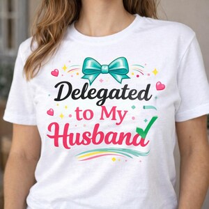 May include: White t-shirt with the text "Delegated to My Husband" in black and pink script. A teal bow and decorative hearts and stars embellish the design. The shirt is a casual wear item.