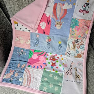 May include: A patchwork baby blanket with a pink fleece backing and trim. The front features various fabric squares with whimsical designs, including a hot air balloon, bunnies, giraffes, and the text "my first christmas".
