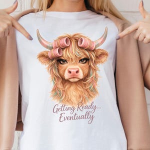 Puede incluir: Camiseta blanca con una ilustración caprichosa de una vaca de las Tierras Altas con rulos rosas, largas pestañas y pelo colorido. El texto "Getting Ready... Eventually" está debajo. Una prenda de vestir informal.
