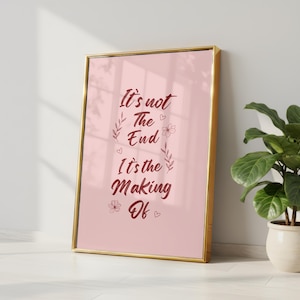 Könnte beinhalten: Gerahmter Druck mit goldenem Rahmen und rosa Hintergrund. Der Druck zeigt das Zitat "It's not The End It's the Making Of" in einer burgunderfarbenen Schriftart mit floralen und herzförmigen Akzenten. Der Druck lehnt an einer weißen Wand.