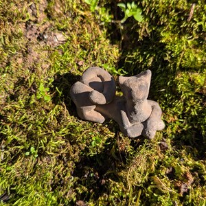 May include: A small, brown, hand-carved cat figurine. The cat is curled up, with its tail wrapped around its body. The figurine is resting on a bed of green moss, with sunlight casting shadows.