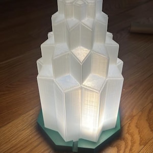 May include: A white, geometric lamp with a layered design, illuminated from within. The lamp sits on a green, hexagonal base. The lamp's design is reminiscent of an art deco skyscraper.