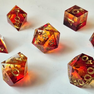 May include: A collection of seven multi-sided dice in various shapes, including a pyramid, cube, and other polyhedrons. The dice are transparent with a fiery red and orange colour scheme, and gold-coloured numbers. Suitable for tabletop gaming.