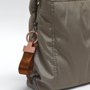May include: A taupe-coloured nylon handbag featuring a smooth, padded texture with visible stitching along the edges. A silver-toned zip runs along the side, attached to a metallic rose-gold ring holding a short, textured bronze-coloured fabric strap.