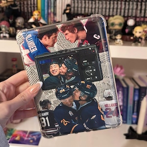 Peut inclure: Coque de téléphone transparente avec un collage de joueurs de hockey s'embrassant, sur fond de journal. La coque présente des images de joueurs en maillots avec des numéros et des noms. La coque est tenue devant une étagère avec des livres et des figurines.
