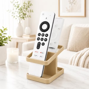 May include: A tan plastic remote control holder with two slots, currently holding two white television remote controls. The front remote features a black circular navigation pad, volume and channel buttons, and logos for Netflix, YouTube, Prime Video, and Disney+.