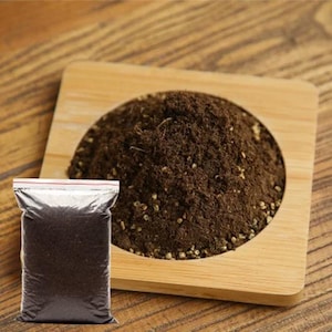 May include: A clear plastic bag filled with dark brown granules, likely spices or herbs, sits in front of a wooden tray holding a larger quantity of the same material. The tray is square with a circular indentation.