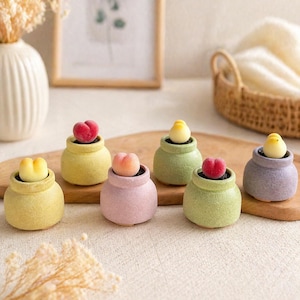 May include: A collection of small, pastel-colored ceramic pots, each containing a miniature fruit-shaped decoration. The pots are in shades of yellow, pink, green, and purple. The fruit decorations include peaches and other yellow fruits. The arrangement is on a wooden surface.