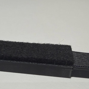 May include: A black cleaning tool featuring a rectangular strip of dense, fuzzy black fabric attached to a rigid, textured black plastic base. The tool has a linear design with a distinct contrast between the soft, fibrous texture and the hard plastic.