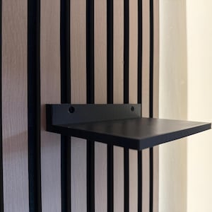 Könnte beinhalten: Ein schwarzes schwebendes Regal an einer Wand mit vertikalen Holzpaneelen. Das Regal ist rechteckig und scheint aus Holz zu sein. Die Wandverkleidung hat abwechselnd helle und dunkle vertikale Streifen.