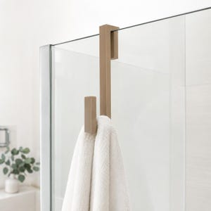 Peut inclure: Un crochet de porte en métal doré mat au design rectangulaire, doté d'un support supérieur plat qui s'adapte solidement sur une paroi de douche en verre. Une serviette blanche est suspendue au crochet inférieur, qui présente une forme rectangulaire.