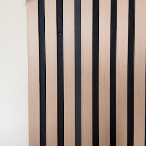 Può includere: Pannello in legno con strisce verticali alternate marrone chiaro e nere. Un cavo USB bianco è collegato al pannello, arcuandosi attraverso il telaio. Il pannello è montato su un muro bianco.