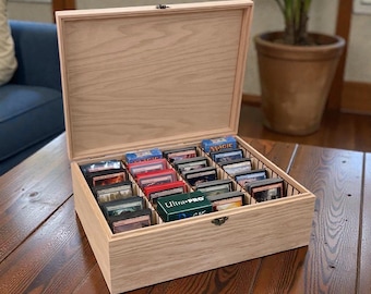 May include: A light-colored wooden storage box with an open hinged lid, featuring internal dividers organized into rows. The box contains multiple stacks of trading cards in protective sleeves, including one green box labeled Ultra-PRO.