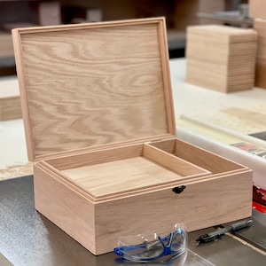 Könnte beinhalten: Geöffnete Holzkiste mit Scharnierdeckel, mit hellfarbiger Holzmaserung. Der Innenraum hat ein Tablett und ein Fach. Ein schwarzer Riegel sichert den Deckel. Schutzbrille und Stift sind im Vordergrund.