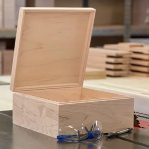 May include: An open wooden box made of light wood with a natural finish. The box is empty and has a lid that is slightly ajar. A pair of safety glasses and a pen are lying on a surface in front of the box.