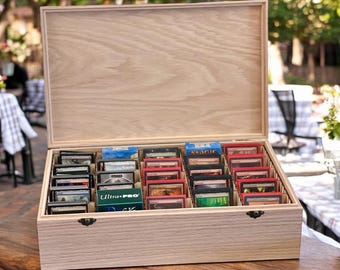 May include: A rectangular light-colored wooden storage box with an open hinged lid, featuring multiple internal dividers filled with rows of trading cards in protective sleeves, including one green box labeled Ultra-PRO.