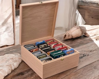 May include: A light-colored wooden storage box with an open hinged lid, featuring internal dividers that organize multiple stacks of trading cards. The box includes a small metal latch on the front and contains various card decks, including one labeled Ultra Pro.