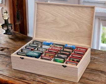 May include: A light-colored wooden storage box with an open hinged lid, featuring multiple internal grid compartments filled with various trading card decks in protective sleeves, including a blue box labeled Ultra PRO.